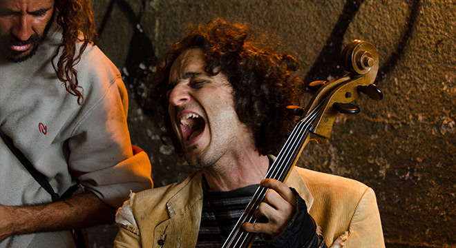 Imagem Feverestival 1 Na foto dois artistas, um a esquerda de pé, olhando para o outro ao centro mais abaixo, tocando violoncelo, expressão do rosto com os olhos fechados, boca aberta e a cabeça levemente inclinada para trás, ao fundo parede com textura, ambiente com pouca luz.