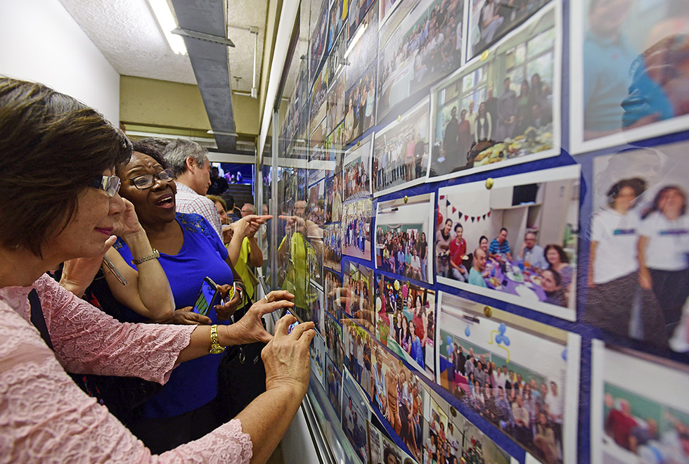 Mulher em primeiro plano tira foto de mural de fotos. Outras pessoas ao redor acompanham e observam o mural