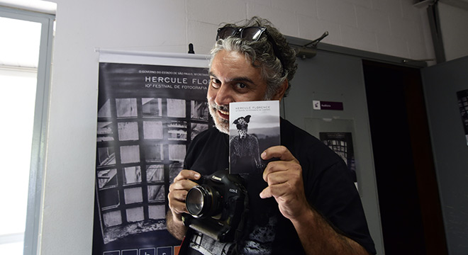 homem com camera em uma mão e folder na outra em frente so cartaz com festival