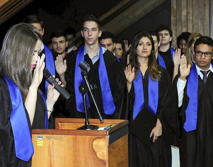 Imagem mostram diversos alunos, com as mãos levantadas, em momento de juramento da formatura do ProFIS 2015. Todos os formandos estão com becas pretas e faixas azuis