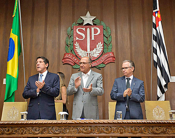 Autoridades em pé batendo palmas, bandeiras hasteadas, do lado direito bandeira do estado de São Paulo, do lado esquerdo bandeira do Brasil, ao fundo em parede de madeira com o brasão do estado de São Paulo