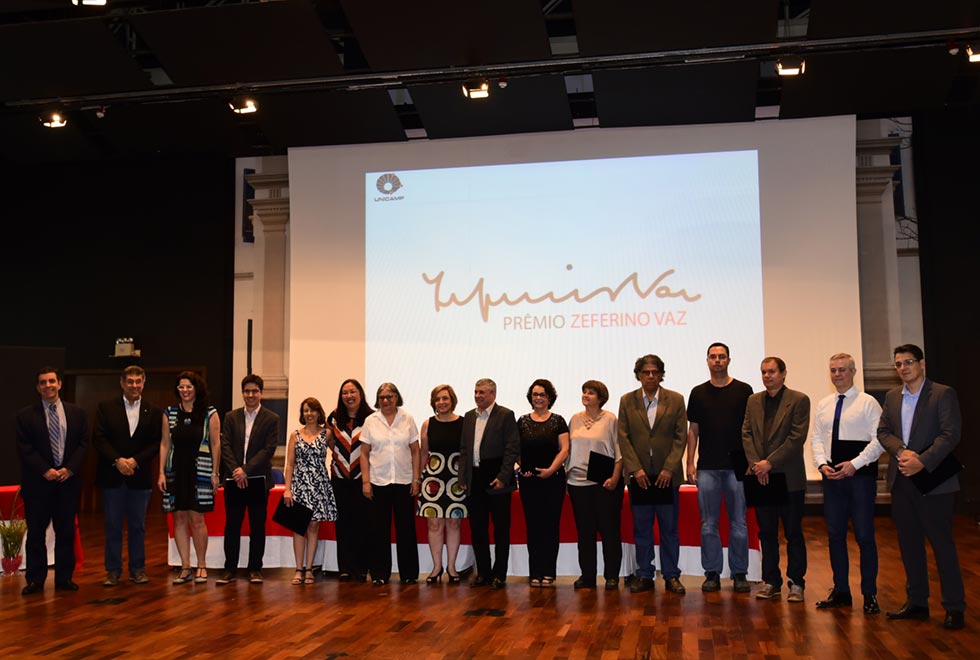 Em imagem frontal, em ambiente com pouca luz, no palco de um grande auditório com assoalho de madeira, dezesseis pessoas, entre homens e mulheres, em pé, perfiladas, uma ao lado da outra, a maioria sorrindo, sendo que alguns têm nas mãos pastas pretas. A maioria veste roupa social como blazer, vestido e camisa de botões. Às costas deles, ao fundo, há uma grande tele de projeção de data show onde se lê Prêmio Zeferino Vaz, e acima dele, a assinatura de Zeferino Vaz.