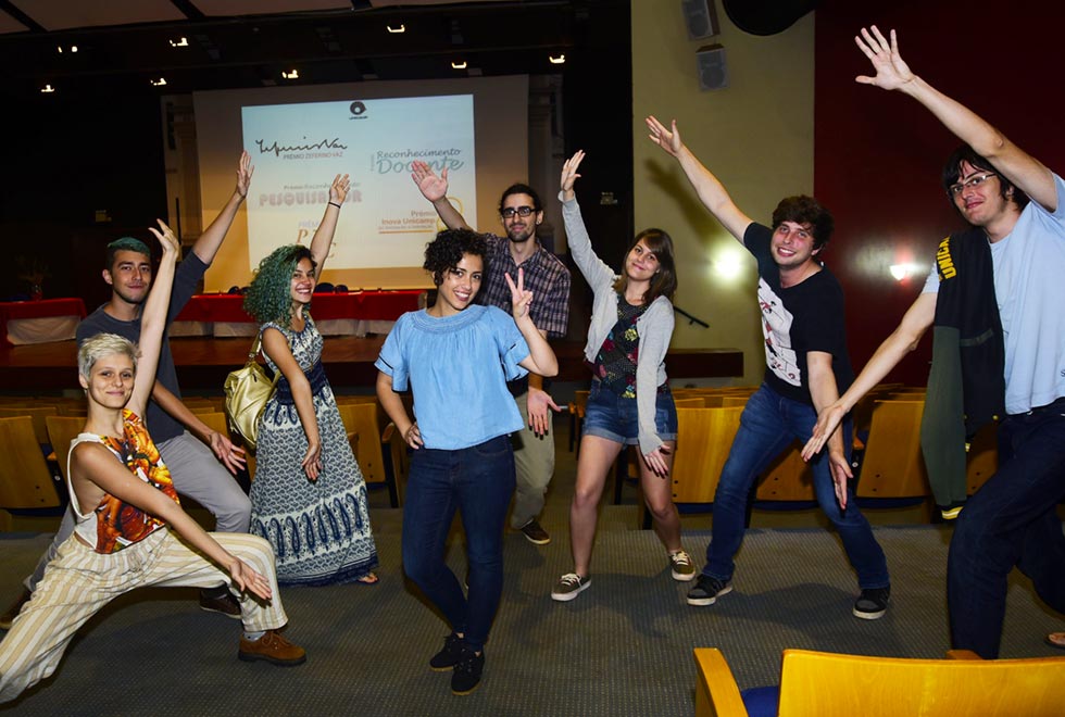 Em imagem frontal, em ambiente com pouca luz, em amplo auditório, grupo de sete jovens, em pé, fazendo pose colocando um braço estendido para cima e ou outro para baixo; e uma perna para frente e outra para trás, posicionados ao redor de uma outra jovem, em pé, ao centro na imagem. A jovem ao centro está com mão direita na cintura, e faz um sinal de paz e amor com a mão esquerda. Todos vestem roupas casuais e estão sorrindo. Ao fundo, às costas deles, estão o palco do auditório e uma ampla tela de data show