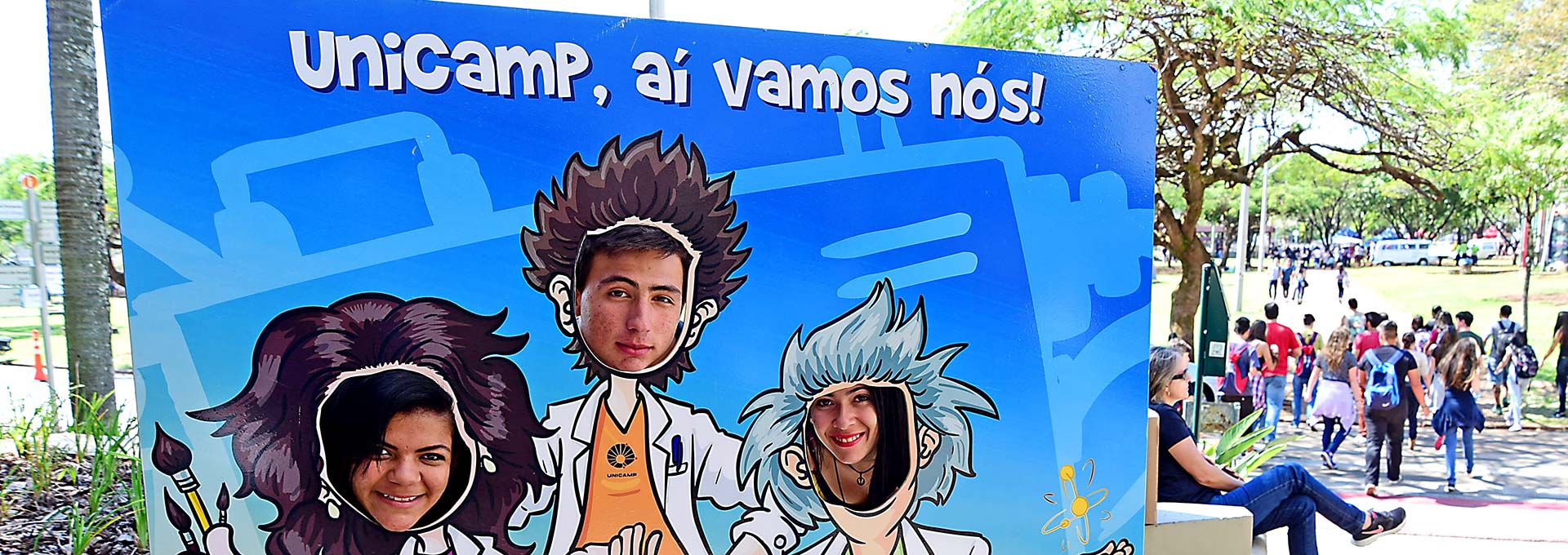 Participantes se divertem durante uma das edições da UPA | Foto: Antonio Scarpinetti Em foco à esquerda, painel com a frase "Unicamp, aí vamos nós!" e com imagem de três pessoas vestindo jaleco onde os visitantes posicionaram seus rostos num fundo vazado para a foto; à direita, desfocados, estão visitantes passeando pelo campus.