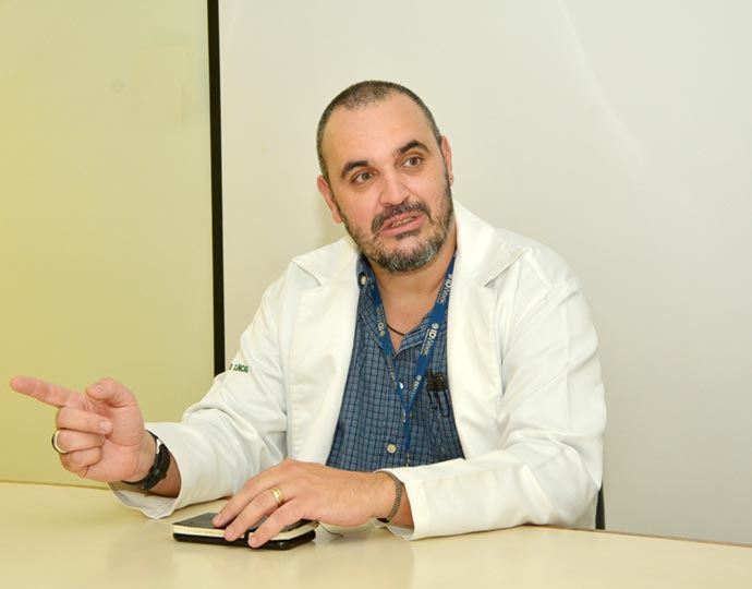 Rodrigo Angerami, médico infectologista da Seção de Epidemiologia Hospitalar do HCs da Unicamp Audiodescrição: Em sala, em imagem frontal, homem sentado em cadeira, sendo que mantém o braço esquerdo apoiado sobre uma mesa de madeira que encontra-se à frente dele, segurando com a mão uma pequena agenda e um telefone celular, enquanto que, com a mão direita, gesticula apontando o dedo indicador para a esquerda na imagem. Ele veste um jaleco branco sobre camisa social verde. Imagem 1 de 1.