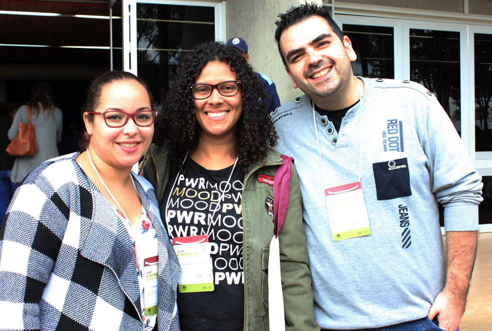 Marta Souza (à esq.), Mariana Donadeli e Tiago de Campos: conteúdo da oficina auxilia no trabalho com os alunos Audiodescrição: Em área externa, imagem frontal e em plano médio, três pessoas em pé, abraçadas, uma ao lado da outra, sendo que todos sorriem e olham para as lentes da câmera fotográfica. Eles usam crachá no pescoço, e blusas de mangas compridas. Ao fundo, às costas deles, a entrada de uma edificação, com portas de vidro transparente e estruturas de alumínio branco. Imagem 3 de 3.