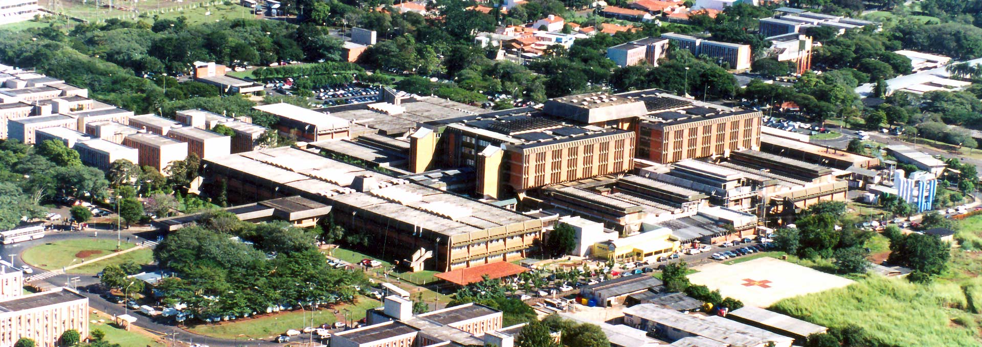 Imagem aérea do Hospital de Clínicas da Unicamp Audiodescrição: Imagem panorâmica aérea de amplo complexo hospitalar constituído de um conjunto de prédios, mas todos com poucos andares. No entorno das edificações, há uma ampla área com vegetação e muitas árvores, além de outras edificação também de baixa altura. O dia está ensolarado. Imagem 1 de 1.