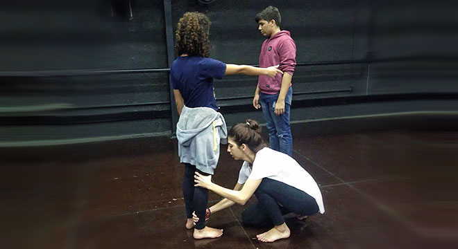 Alunos do curso Exato realizam atividade de dramaturgia. Na foto três alunos aparecem. Dois estão em pé e um terceiro abaixado.