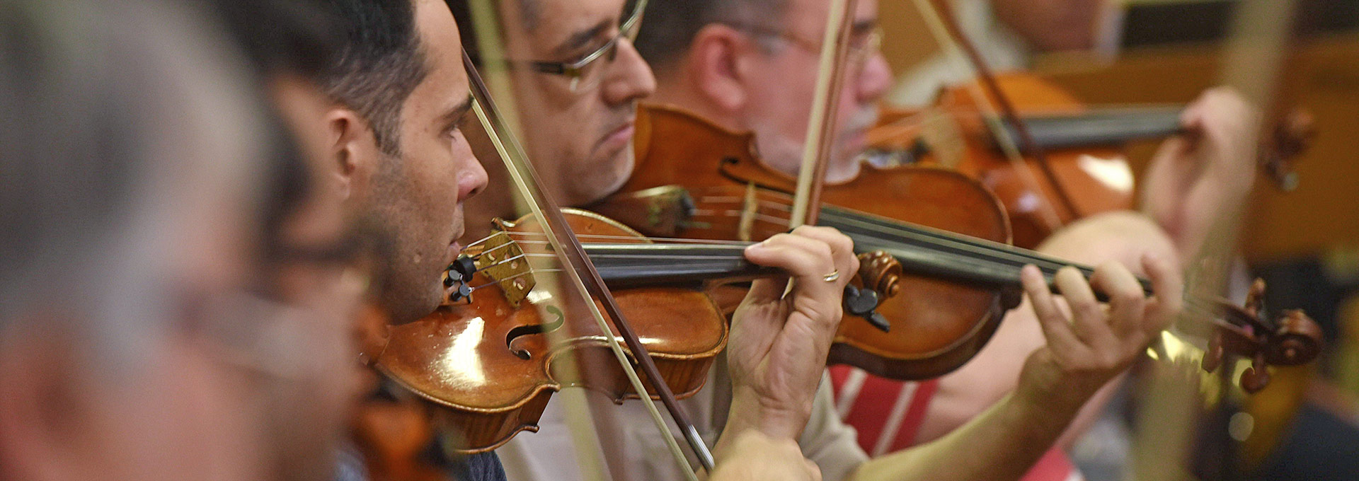 Audiodescrição: Em área interna, imagem close-up e em perspectiva, três homens tocam violino, dispostos um ao lado do outro, sendo que estão posicionados da esquerda para a direita, com olhares voltados para a direita. Imagem 1 de 1.