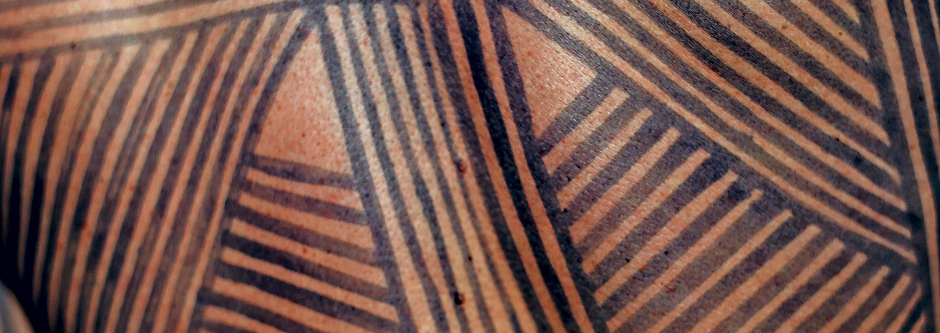Audiodescrição: Em imagem close-up e frontal, pintura indígena brasileira feita na pele de uma pessoa com uso de tinta preta e elaborada com traços verticais e na diagonal, unidos, como que formando triângulos. Imagem 1 de 1.
