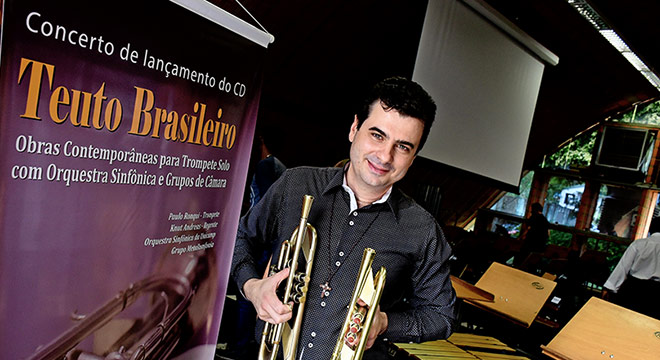 O trompetista Paulo Ronqui aparece ao lado do cartaz do evento segurando dois instrumentos de sopro