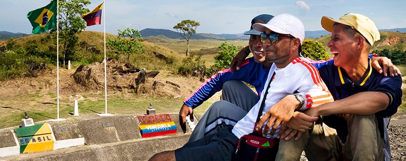 Audiodescrição: Em área externa, imagem em perspectiva, três homens sentados sobre uma pedra, à direita na imagem, abraçados e sorrindo, com olhares voltados para a esquerda da imagem. Todos usam bonés e camisetas. Eles estão na divisa entre Venezuela e Brasil. Ao fundo, à esquerda, há duas bandeiras desses países hasteadas em mastros, com montanhas ao fundo. Junto às bandeiras, abaixo delas, há um paredão de concreto com duas pinturas em alusão às bandeiras de Brasil e Venezuela. Imagem 1 de 1.