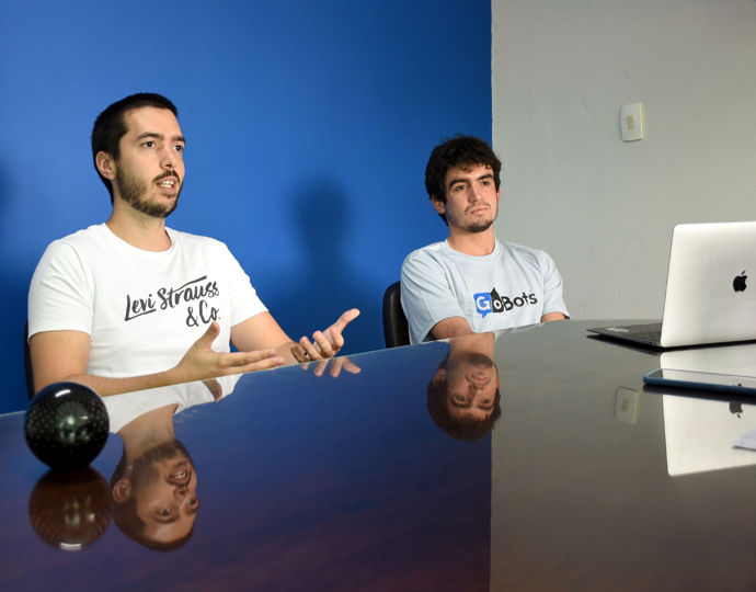 Foto mostra, da esquerda para a direita, Samuel Birocchi e Victor Hochgreb. Eles estão sentados à mesa com um laptop à frente