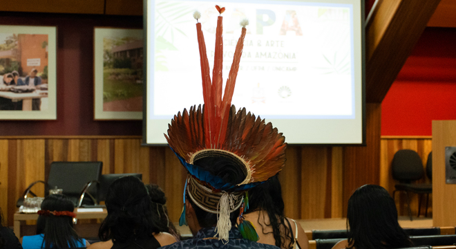 foto mostra aluno indígena sentado na plateia usando um cocar