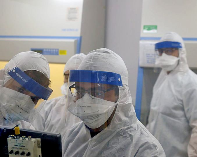 Pesquisadores com roupas brancas e viseiras especiais para manipulação do vírus em laboratório  