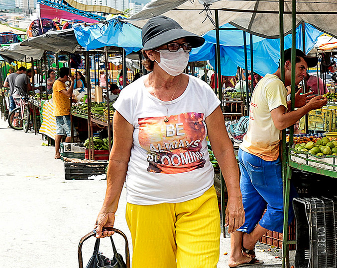foto mostra senhora em uma feira livre usando máscara de proteção. ela usa óculos, chapéu e puxa um carrinho de compras. clique enter para acessar
