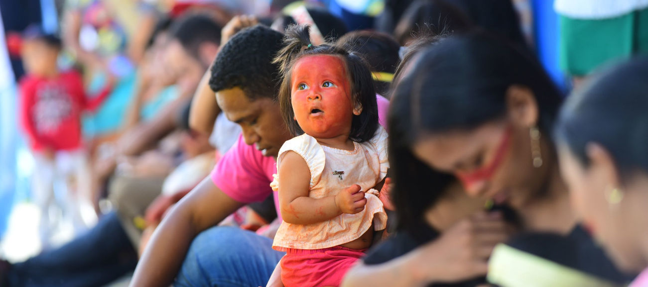 audiodescrição: fotografia colorida focada em rosto de criança indígena pintado de urucum; ela está no colo da mãe e há mais mulheres sentadas ao lado