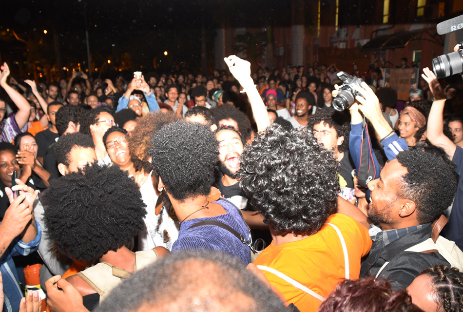 Em área externa, a noite, grupo de mais de 100 pessoas, em pé, comemora sorrindo e erguendo os braços