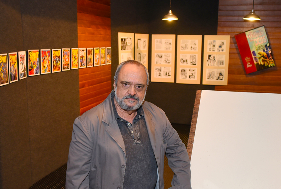 Homem em pé, ao centro, vestindo blazer e camisa social em tom escuro, tendo às costas, à esquerda, fixado em parede de madeira, várias capas coloridas e em preto e branco de páginas com ilustrações de histórias em quadrinhos