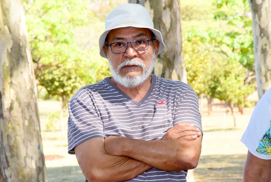 Ao ar livre, em ampla praça gramada com várias árvores altas ao fundo, homem, em pé, de braços cruzados, sendo que ele usa óculos e veste camisa lilás e um chapéu de pescador branco. Imagem 5 de 8.