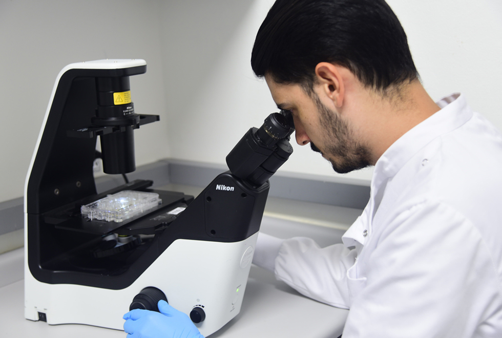 Em laboratório, imagem de perfil e em close-up, homem observa material por meio do visor um grande microscópio que se encontra sobre uma bancada, sendo que ele veste um jaleco branco. Imagem 3 de 5.