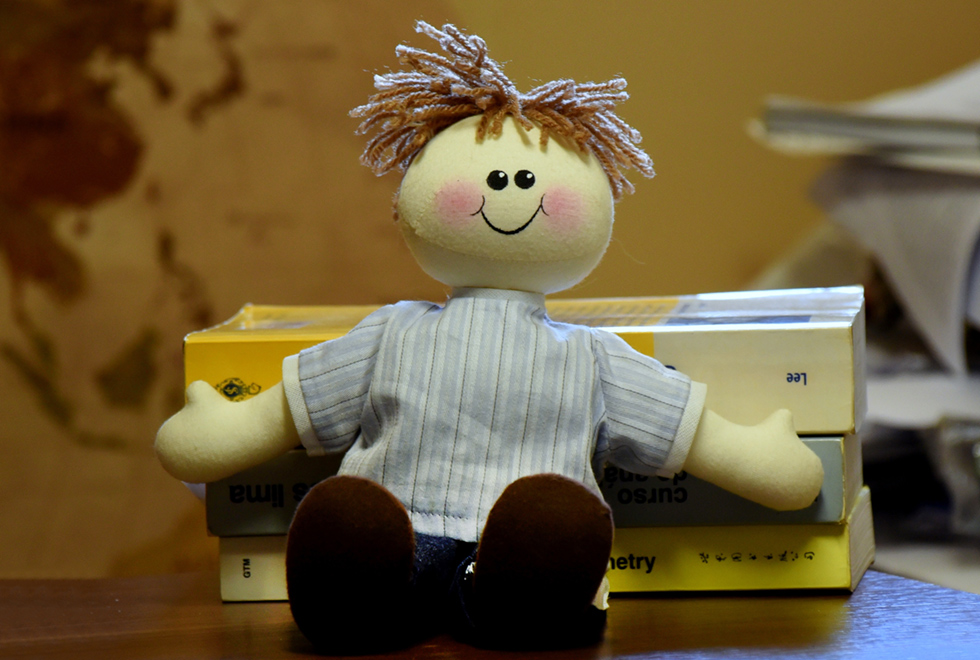 Imagem frontal de um boneco de pano no formato de um menino, sentado e apoiado sobre uma mesa da madeira. O boneco tem cerca de trinta centímetros de altura, veste uma blusa azul listrada e está sorrindo. Imagem 4 de 4.