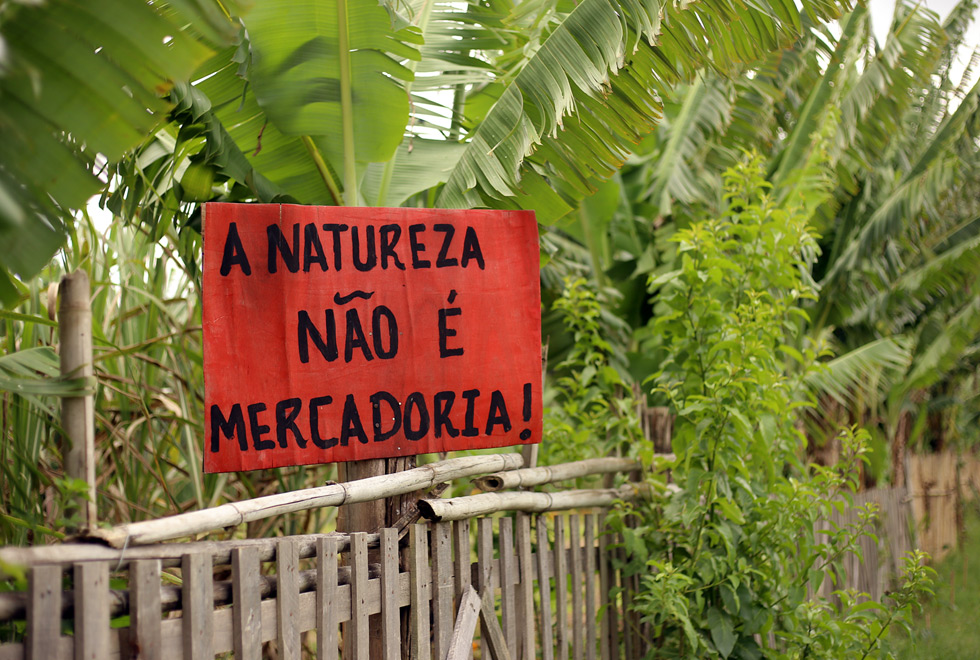 audiodescrição: fotografia colorida de placa vermelha com dizeres em preto escrito "natureza não é mercadoria"