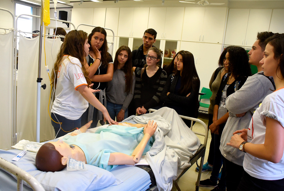 foto mostra estudantes visitando um laboratório de medicina com um boneco deitado em cama hospitalar