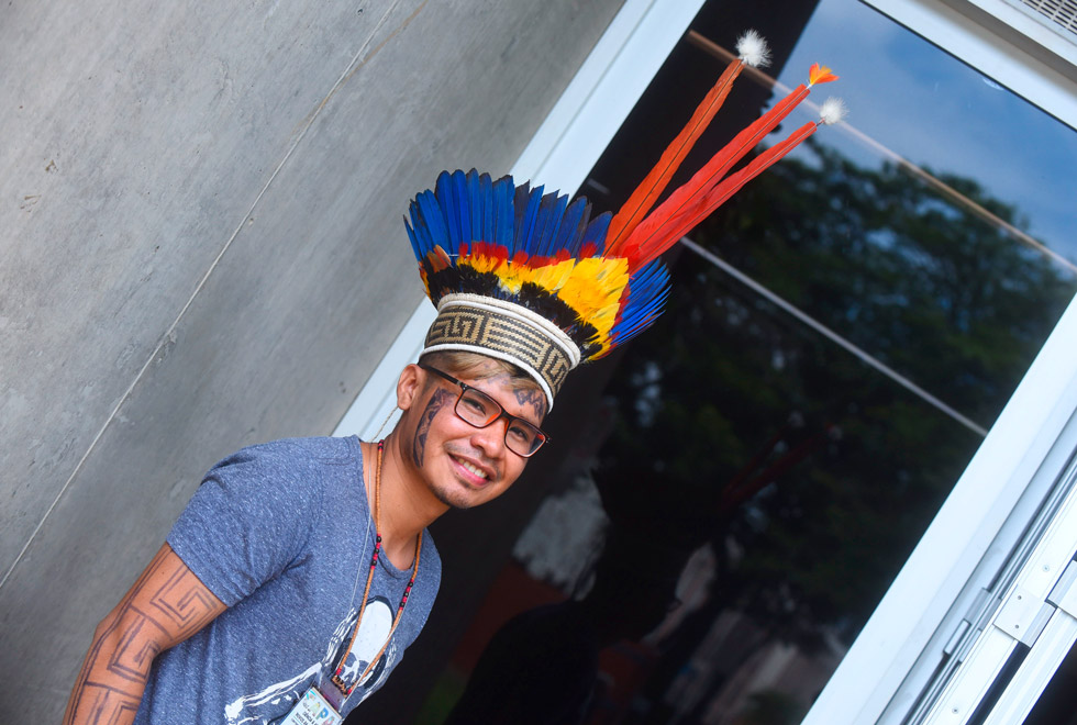 audiodescrição: fotografia colorida. jovem indígena, com cocar de cores amarelo, azul e laranja, sorri para a foto. ele tem pinturas indígenas no rosto e nos braços e usa óculos.