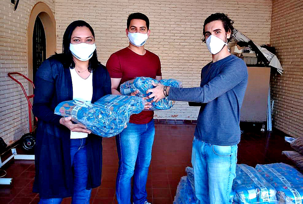 foto mostra aluno, à direita, entregando alimentos embalados a dois representantes de uma entidade beneficente. os 3 usam máscaras