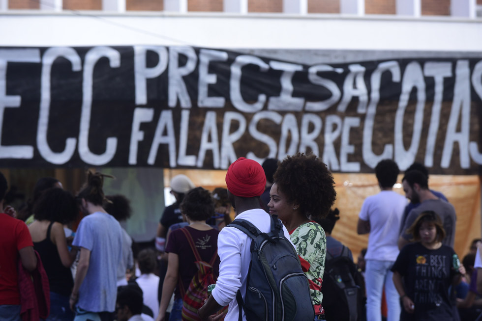 audiodescrição: fotografia colorida mostra estudantes negros e ao fundo uma faixa com os dizeres "precisamos falar sobre cotas"