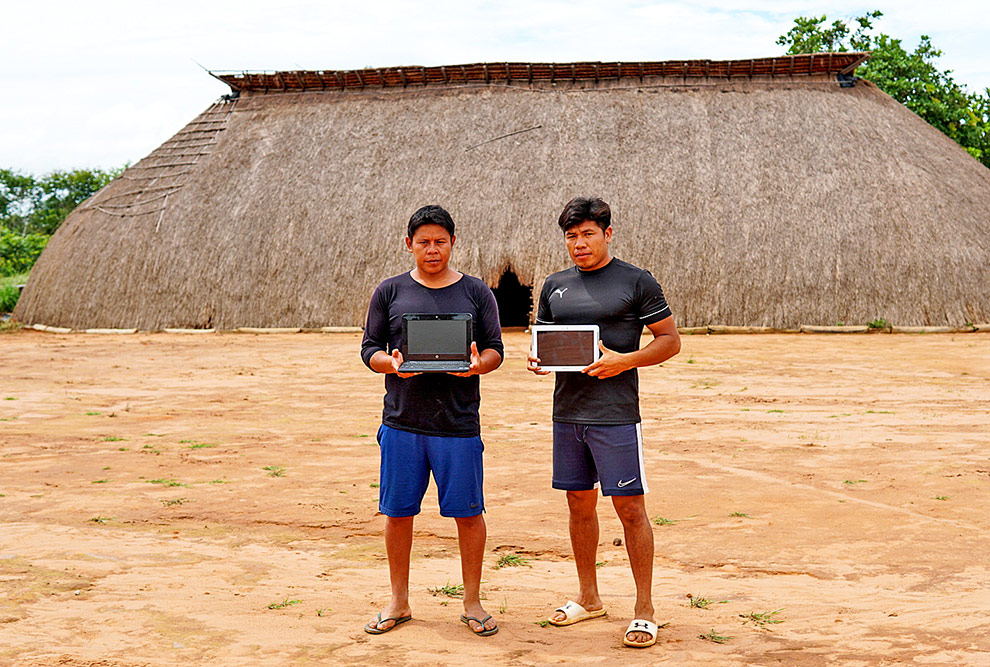 audiodescrição: fotografia colorida mostra dois estudantes indígenas, em frente a uma oca, segurando um computador e um tablet; eles estão posando para a foto
