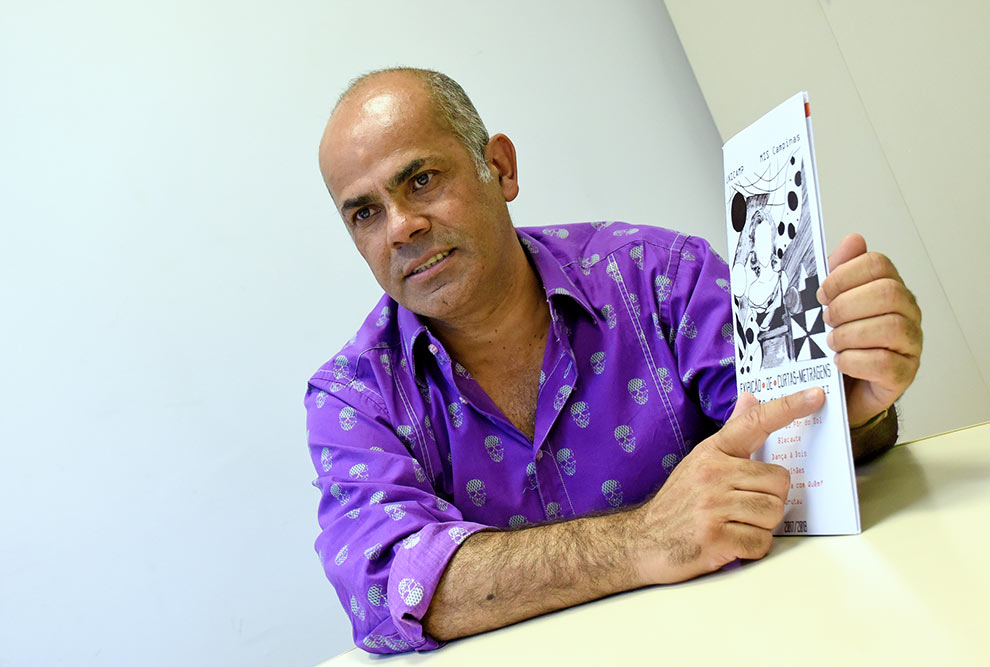 Noel Carvalho aparece sentado segurando o cartaz da mostra do ano passado. 