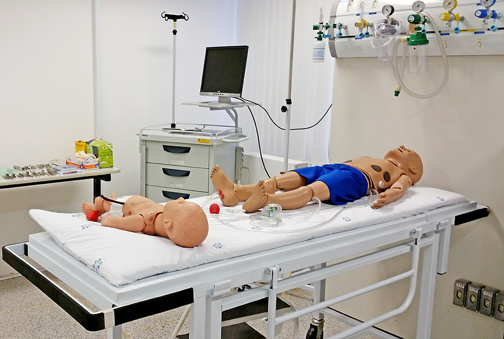 imagem mostra sala com manequins usados para o ensino. Há uma maca com bonecos de duas crianças e equipamentos de emergencia