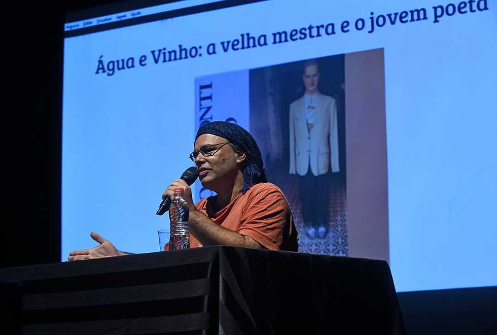 Renato está no auditório do IA, sentado com microfone na mão. Veste camiseta cor laranja e usa um lenço azul nos cabelos