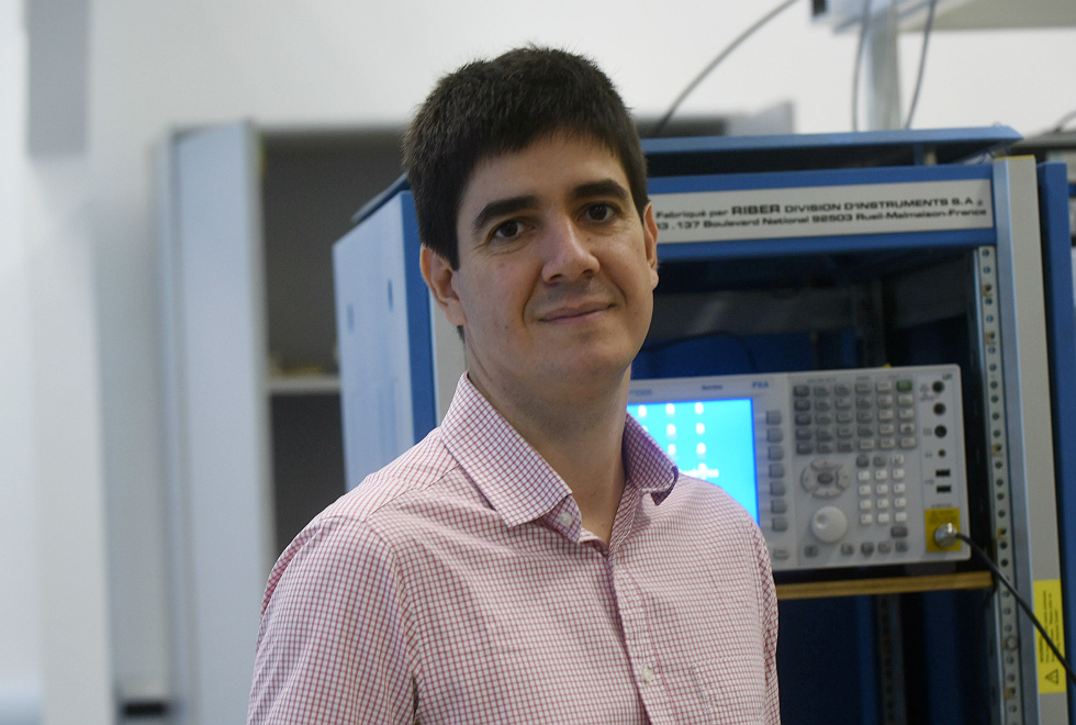 O professor Gustavo olha para a camera, ele está de camisa rosa xadrez em frente aos equipamentos do laboratório