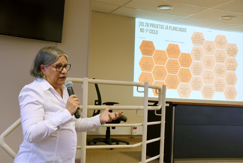 Professora Teresa Atvars faz apresentação do projeto de gestão estratégica da universidade. Ela está no lado esquerdo da foto, olhado em direção ao lado direito, com um microfone na mão. Ao fundo, uma tela apresenta uma imagem com parte da projeção sobre o tema.