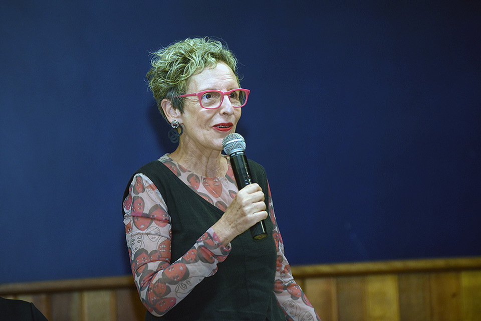 Mulher de cabelos curtos e óculos vermelhos em pé fala com o microfone na mão