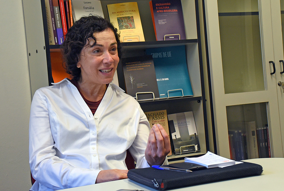 A professora Márcia Abreu, diretora da Editora da Unicamp gesticula durante a entrevista para a matéria. Ela está de camisa branca, sentada em frente a uma prateleira de livros