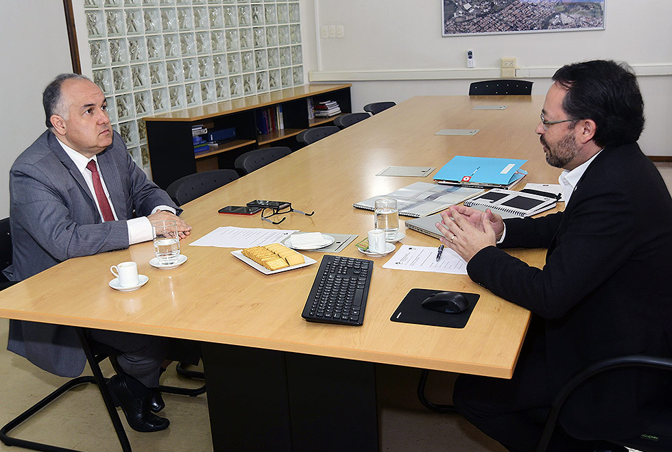 O secretário adjunto Romildo Campello está sentado em frente ao professor Fernando Hashimoto. Ambos vestem roupas mais formais. Sobre a mesa há vários objetos como teclado de computador, café e bolachas