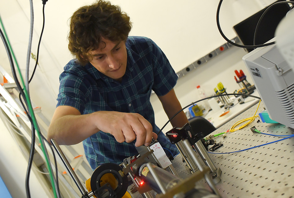 O pesquisador Tiago Alegre, de frente para a câmara, vestindo camisa xadrez de mangas curtas, ajusta esquipamentos eletrônicos em bancada de seu laboratório