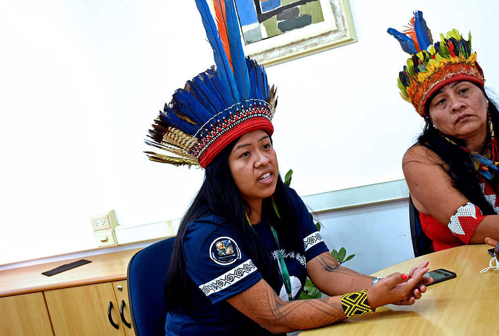 Foto de duas mulheres indígenas. Elas estão sentadas e usam cocar, colares de contas e enfeites nos braços. 