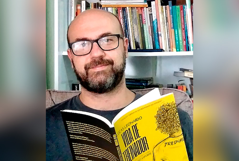 Foto de um homem branco. Ele usa barba e bigode pretos, óculos, camiseta escura e está segurando um livro. Atrás dele há uma estante com livros.