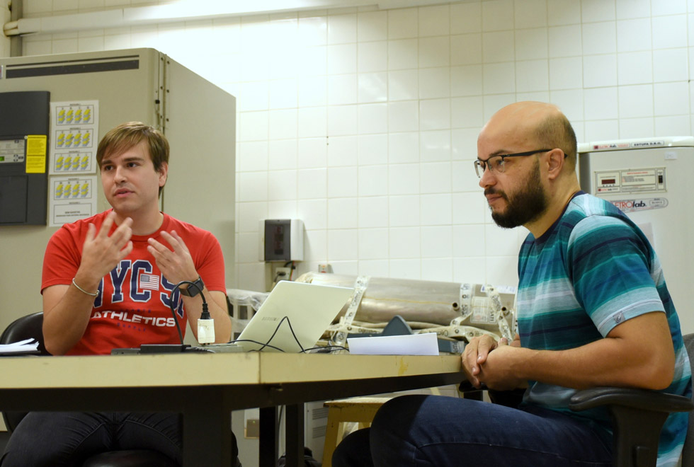 audiodescrição: fotografia colorida mostra dois homens, estudante e professor, em torno de uma mesa. o mais jovem, o estudante, gesticula enquanto fala. eles estão em um laboratório. 