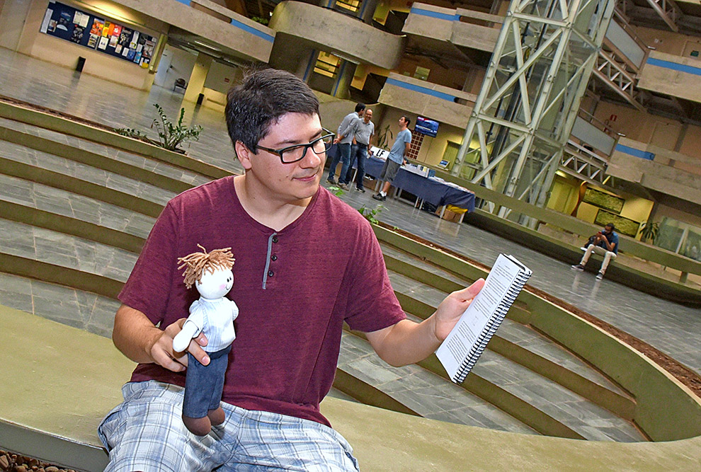 Régis Varão brinca, mostrando um livro para o boneco de pano. Ele está sentado em uma mureta no pátio interno do instituto de matemática