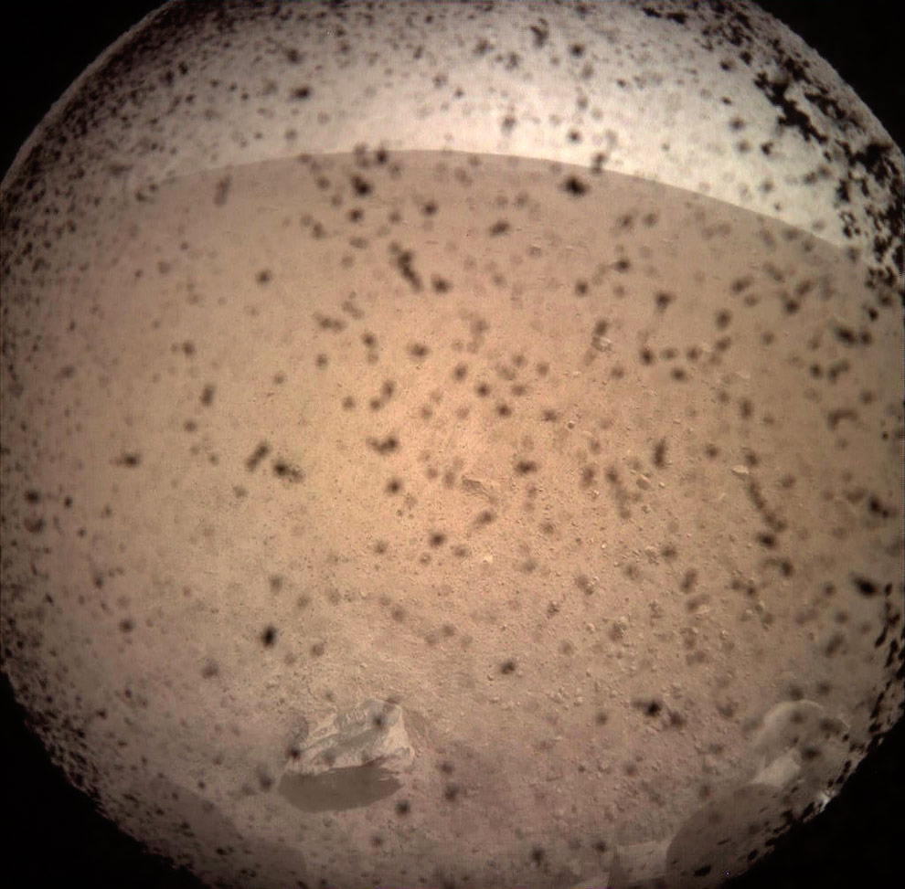Primeira imagem transmitida pela InSight. As manchas são poeira mas pode-se observar parte do trem de pouso no canto inferior direito.(Crédito: NASA/JPL-Caltech)