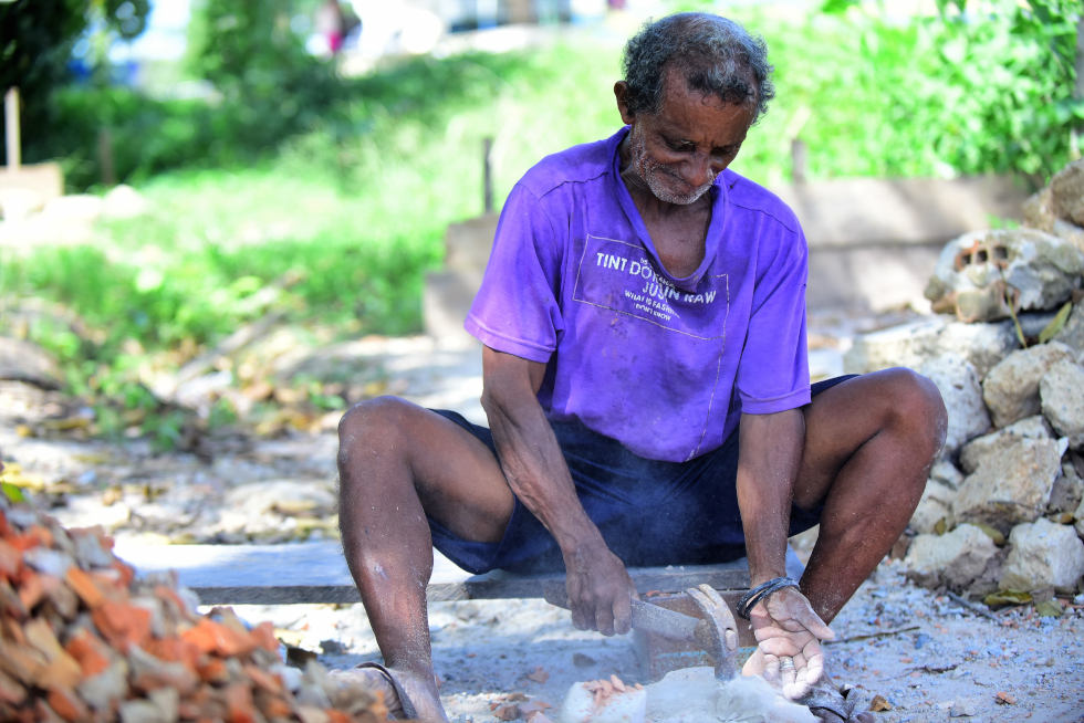 audiodescrição: fotografia colorida mostra trabalhador sentado quebrando pedras com um martelo