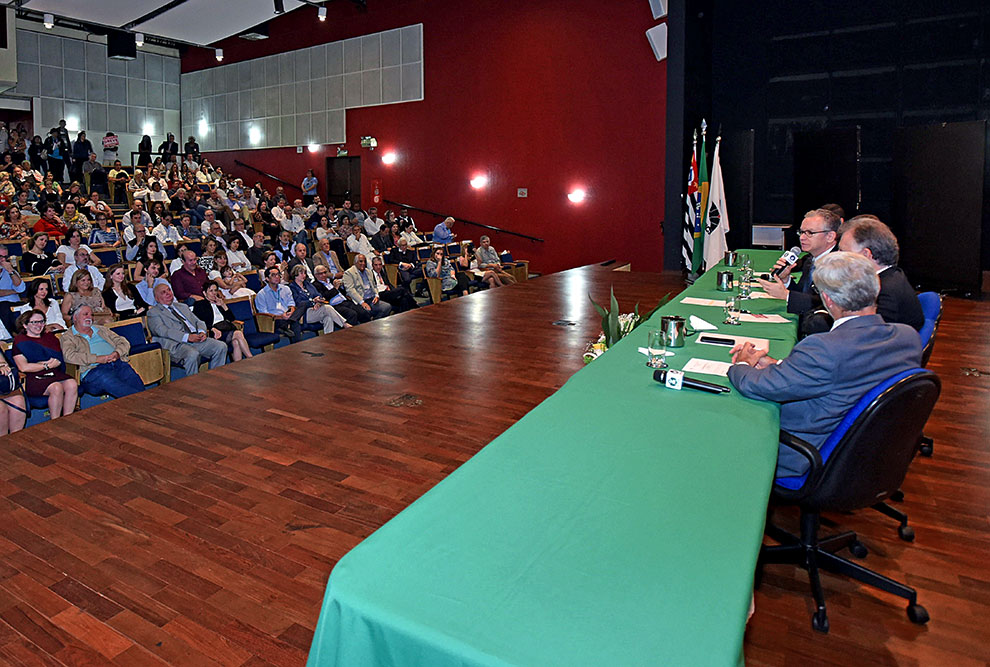 Imagem aberta mostra o auditório cheio e a mesa diretora da solenidade no momento em que fala o reitor Marcelo Knobel