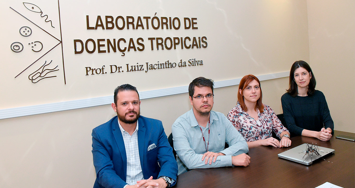 Sentados em volta de uma mesa de madeira, Fabio,rosto com barba, vestindo paletó azul e camisa azul clara, mãos entrelaçadas, José, com óculos e barba curta, vestindo camisa azul clara de manga longa, mão esquerda sobre o braço direito, Juliana ,cabelo avermelhado, com camisa estampada e mãos sobre a mesa, Carla, cabelos pretos blusa azul marinho, mãos uma em cima da outra sobre a mesa, atrás uma parede bege, com os dizeres em marrom: Laboratório de Doenças Tropicais Prof. Dr. Luiz Jacintho da Silva