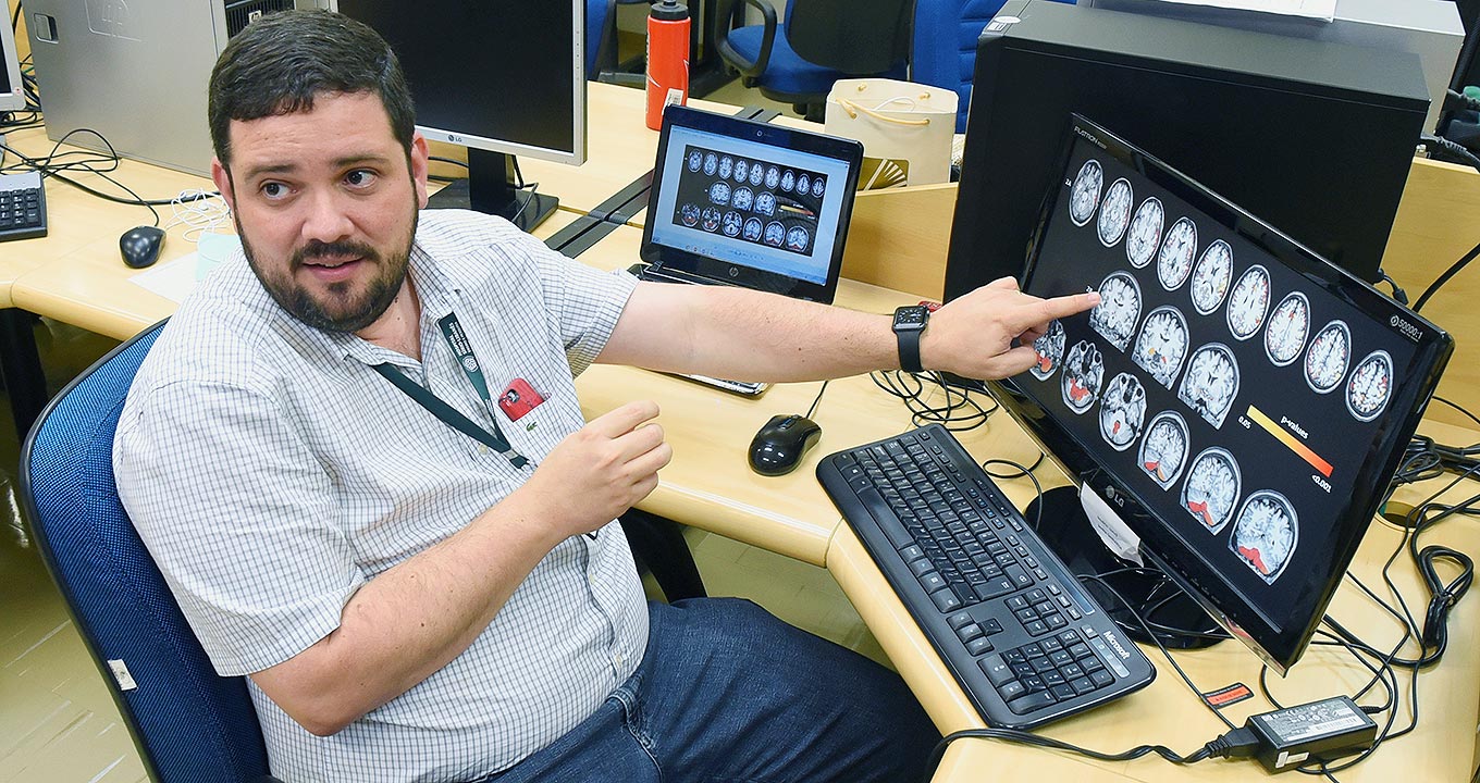 Audiodescrição:Em sala, imagem lateral, homem sentado em cadeira à esquerda na imagem, com olhar voltado à esquerda, aponta com braço esquerdo para monitor de lcd tocando a tela com dedo indicador, enquanto mantém o braço direito erguido e encostado lateralmente na barriga. À frente dele, há uma mesa de madeira em formato l, sobre a qual está esse monitor que exibe dezoito pequenas imagens de cérebros, agrupados um ao lado do outro formando três fileiras. As imagem parecem exame de tomografia. Imagem 1 de 1